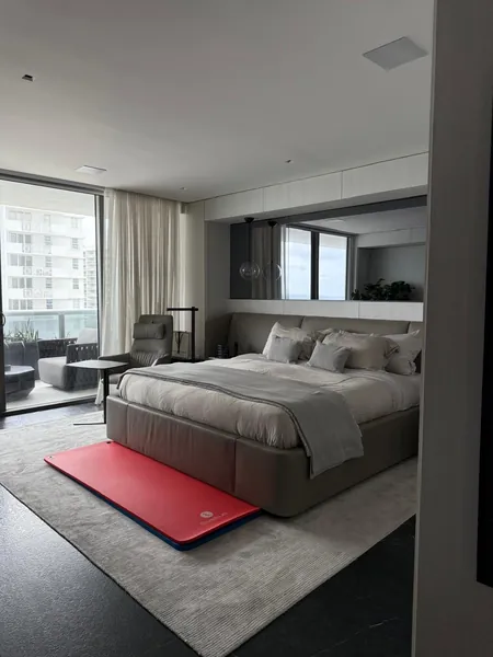 Bedroom with a made bed, pale rug, red exercise mat and windows opening onto a balcony.